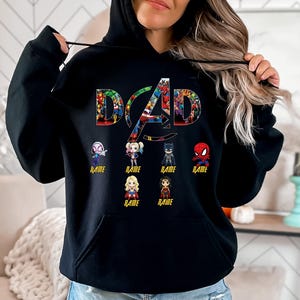 Peut inclure: Un sweat &agrave; capuche noir avec le mot DAD en grandes lettres remplies de divers graphismes de super-h&eacute;ros. Sous le texte se trouvent six petites figurines de super-h&eacute;ros style chibi, chacune avec le mot NAME imprim&eacute; en jaune en dessous.