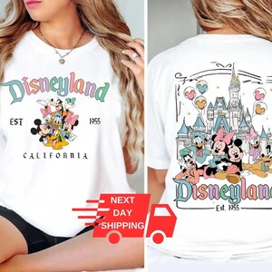 Puede incluir: Camiseta blanca con el logotipo de Disneyland en colores pastel y un gr&aacute;fico de personajes de Disney. La parte trasera de la camiseta muestra un castillo y m&aacute;s personajes. El texto incluye "EST 1955" y "CALIFORNIA".