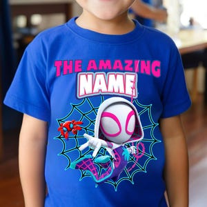 Op de afbeelding: Blauw t-shirt met een superheldenafbeelding. Het ontwerp bevat een personage met een wit en roze masker, een spinnenweb en de tekst "THE AMAZING NAME". Een rode spinnenrobot is ook aanwezig.