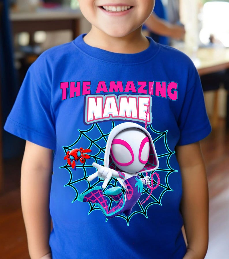 Op de afbeelding: Blauw t-shirt met een superheldenafbeelding. Het ontwerp bevat een personage met een wit en roze masker, een spinnenweb en de tekst "THE AMAZING NAME". Een rode spinnenrobot is ook aanwezig.