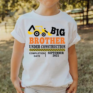 May include: White t-shirt with a construction theme. The shirt features a yellow and black excavator graphic and the words "Big Brother Under Construction" in black and orange. The completion date is September 2025.