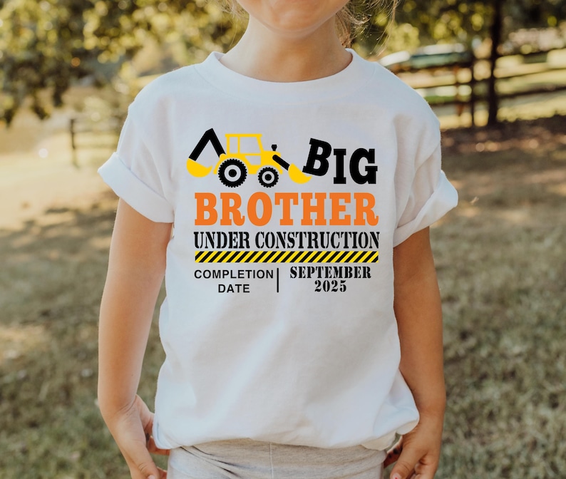 May include: White t-shirt with a construction theme. The shirt features a yellow and black excavator graphic and the words "Big Brother Under Construction" in black and orange. The completion date is September 2025.
