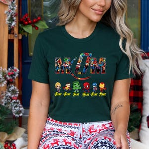 Peut inclure: Un t-shirt &agrave; manches courtes vert fonc&eacute; avec le mot MOM &eacute;crit dans une police de style collage de super-h&eacute;ros color&eacute;s. Sous le texte se trouvent six petites figurines de super-h&eacute;ros avec le mot Name imprim&eacute; en jaune en dessous.