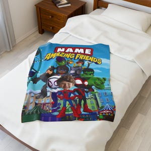 May include: A blue blanket featuring a group of cartoon superheroes with the text "NAME AMAZING FRIENDS". The blanket is decorated with a cityscape and a blue sky background. The blanket is folded on a white bed.