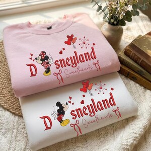 May include: Two sweatshirts, one pink and one white, are stacked. Both feature embroidered designs of Mickey and Minnie Mouse, hearts, and the text "Sneyland Sweethearts" in red. The pink sweatshirt has a Minnie Mouse design, while the white one has a Mickey Mouse design.