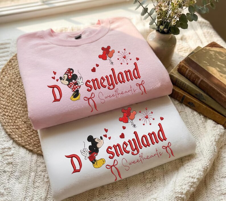 May include: Two sweatshirts, one pink and one white, are stacked. Both feature embroidered designs of Mickey and Minnie Mouse, hearts, and the text "Sneyland Sweethearts" in red. The pink sweatshirt has a Minnie Mouse design, while the white one has a Mickey Mouse design.