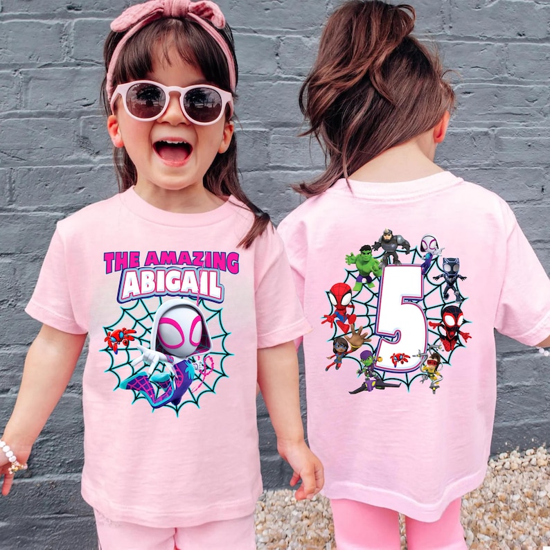 Op de afbeelding: Roze T-shirts met een Spider-Gwen thema. De voorkant heeft de tekst "THE AMAZING ABIGAIL" boven een Spider-Gwen afbeelding in een web. De achterkant toont het cijfer 5 omringd door superhelden.