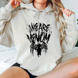 May include: Cream-colored hoodie with the text "WE ARE VENOM" in a black, stylized font, accompanied by a black spider-like graphic. The hoodie features a drawstring hood and long sleeves.