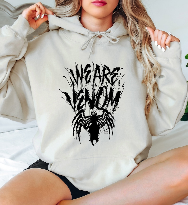 May include: Cream-colored hoodie with the text "WE ARE VENOM" in a black, stylized font, accompanied by a black spider-like graphic. The hoodie features a drawstring hood and long sleeves.