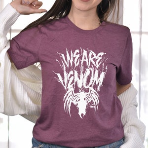 May include: A maroon t-shirt with the text "WE ARE VENOM" in a white, stylized font. Below the text is a white graphic of a spider-like creature. The shirt is short-sleeved and made of a soft material.