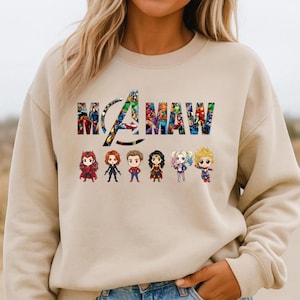 Peut inclure: Un sweat-shirt &agrave; col rond beige avec le texte "MAMA" rempli d'un motif de collage de bandes dessin&eacute;es color&eacute;es. Sous le texte, six petites figurines de personnages de super-h&eacute;ros stylis&eacute;es sont align&eacute;es sur une seule ligne.