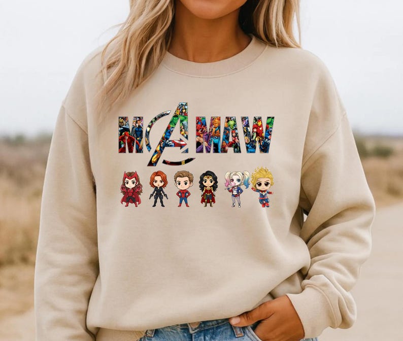 Peut inclure: Un sweat-shirt &agrave; col rond beige avec le texte "MAMA" rempli d'un motif de collage de bandes dessin&eacute;es color&eacute;es. Sous le texte, six petites figurines de personnages de super-h&eacute;ros stylis&eacute;es sont align&eacute;es sur une seule ligne.