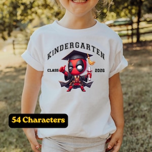 Puede incluir: Una camiseta blanca con un gr&aacute;fico de un personaje rojo y negro con birrete y toga de graduaci&oacute;n, sosteniendo un diploma. El texto dice "KINDERGARTEN" en un arco arriba y "CLASS 2026" a los lados de la ilustraci&oacute;n del personaje.