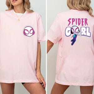 Op de afbeelding: Lichtroze T-shirt met een Spider-Gwen ontwerp. De voorkant van het shirt toont een witte en paarse masker afbeelding. De achterkant van het shirt heeft de woorden "SPIDER GIRL" in paars, met een afbeelding van Spider-Gwen.