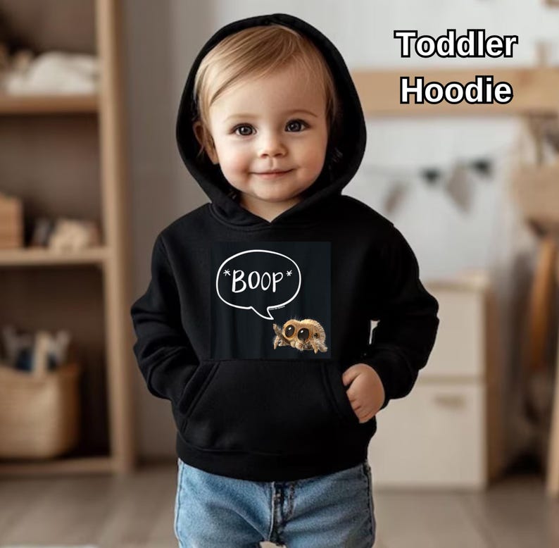 Puede incluir: Sudadera con capucha negra para ni&ntilde;os peque&ntilde;os con bolsillo delantero y capucha. La sudadera con capucha presenta un gr&aacute;fico de una ara&ntilde;a de dibujos animados con la palabra "BOOP" en una burbuja. El texto "Toddler Hoodie" est&aacute; en la esquina superior derecha.