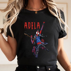May include: Black t-shirt with a red, blue, and black illustration of a cartoon Spider-Man playing a guitar. The text "ADELA" is printed in red above the illustration.