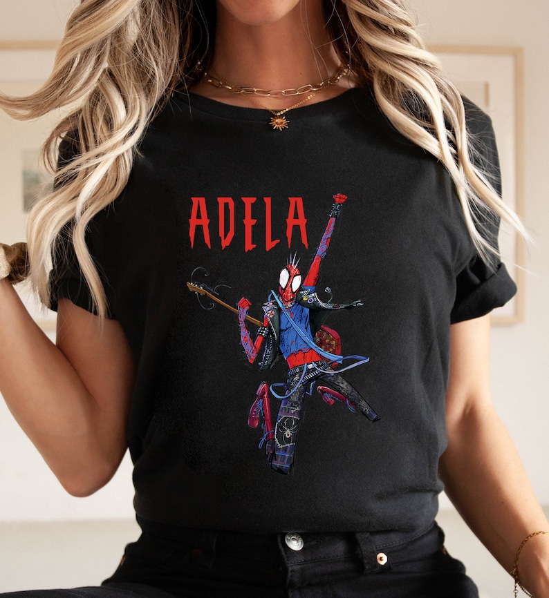 May include: Black t-shirt with a red, blue, and black illustration of a cartoon Spider-Man playing a guitar. The text "ADELA" is printed in red above the illustration.