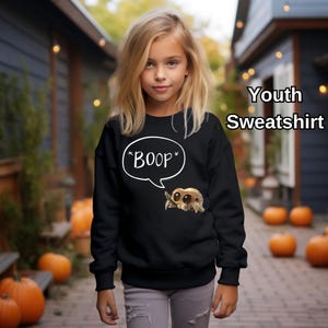 Puede incluir: Sudadera negra juvenil con una ara&ntilde;a de dibujos animados y el texto "*BOOP*" en un bocadillo. La imagen muestra a una persona joven que lleva la sudadera, con calabazas en el fondo.