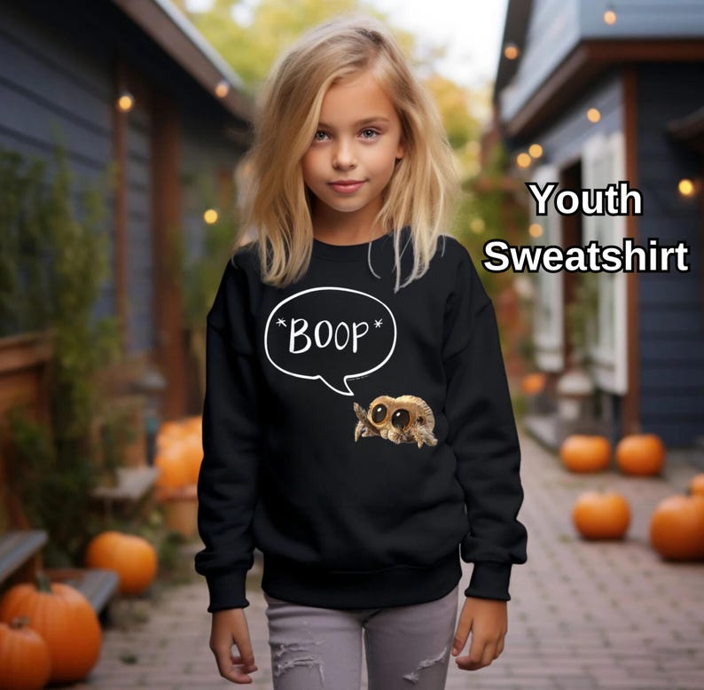 Puede incluir: Sudadera negra juvenil con una ara&ntilde;a de dibujos animados y el texto "*BOOP*" en un bocadillo. La imagen muestra a una persona joven que lleva la sudadera, con calabazas en el fondo.
