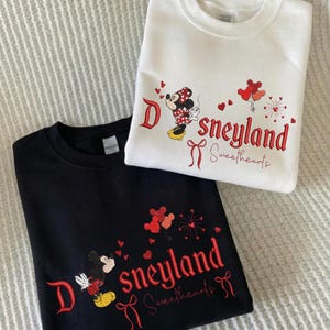 May include: Two sweatshirts, one black and one white, featuring embroidered designs. The designs include the word "Disneyland Sweethearts" in red script, along with images of Mickey and Minnie Mouse. The white sweatshirt has a Minnie Mouse design.