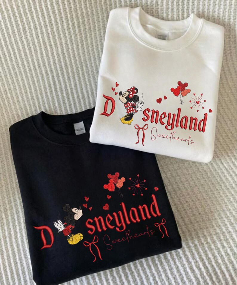 May include: Two sweatshirts, one black and one white, featuring embroidered designs. The designs include the word "Disneyland Sweethearts" in red script, along with images of Mickey and Minnie Mouse. The white sweatshirt has a Minnie Mouse design.