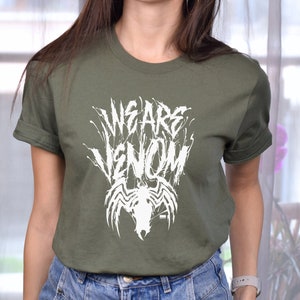 May include: Olive green t-shirt with the text "WE ARE VENOM" in a distressed white font, along with a white spider emblem. The shirt has short sleeves and a crew neck.