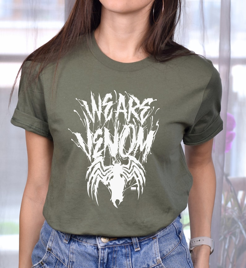 May include: Olive green t-shirt with the text "WE ARE VENOM" in a distressed white font, along with a white spider emblem. The shirt has short sleeves and a crew neck.