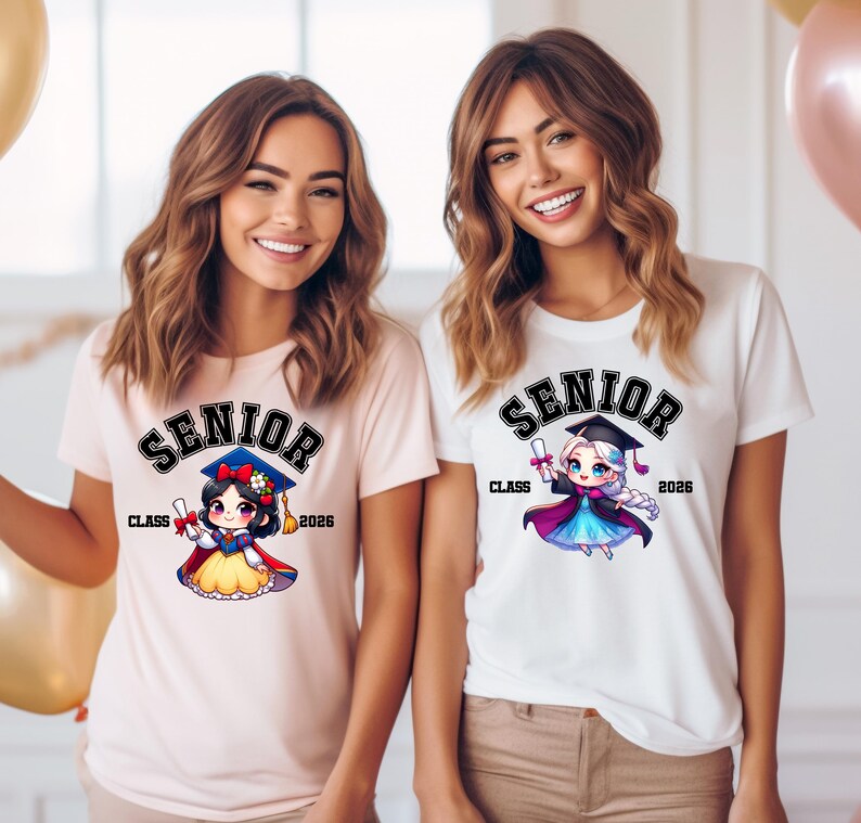 Puede incluir: Una camiseta rosa claro y una camiseta blanca, cada una con un gr&aacute;fico de un personaje de dibujos animados con birrete y toga de graduaci&oacute;n. El texto SENIOR CLASS 2026 est&aacute; impreso sobre y al lado de los personajes en fuente negra negrita.