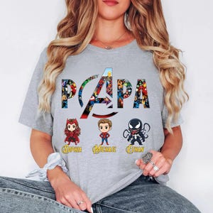 Peut inclure: Un t-shirt gris clair avec le mot PAPA en grandes lettres remplies de divers personnages de super-h&eacute;ros. Sous le texte se trouvent trois figurines de style chibi repr&eacute;sentant Scarlet Witch, Spider-Man et Venom avec les noms Sophia, Michael et Ethan.