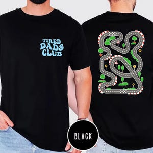May include: Black t-shirt with the text "TIRED DADS CLUB" in light blue on the front. The back of the shirt features a colorful race track design with trees, road signs, and a checkered flag. The shirt is a crew neck.