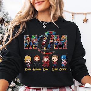 Peut inclure: Un sweat-shirt noir &agrave; manches longues avec le mot MOM en grandes lettres remplies de divers personnages de super-h&eacute;ros. Sous le texte se trouvent cinq figurines de super-h&eacute;ros style chibi avec les noms Alison, Elizabeth, Ferla, Fedro et Freston.