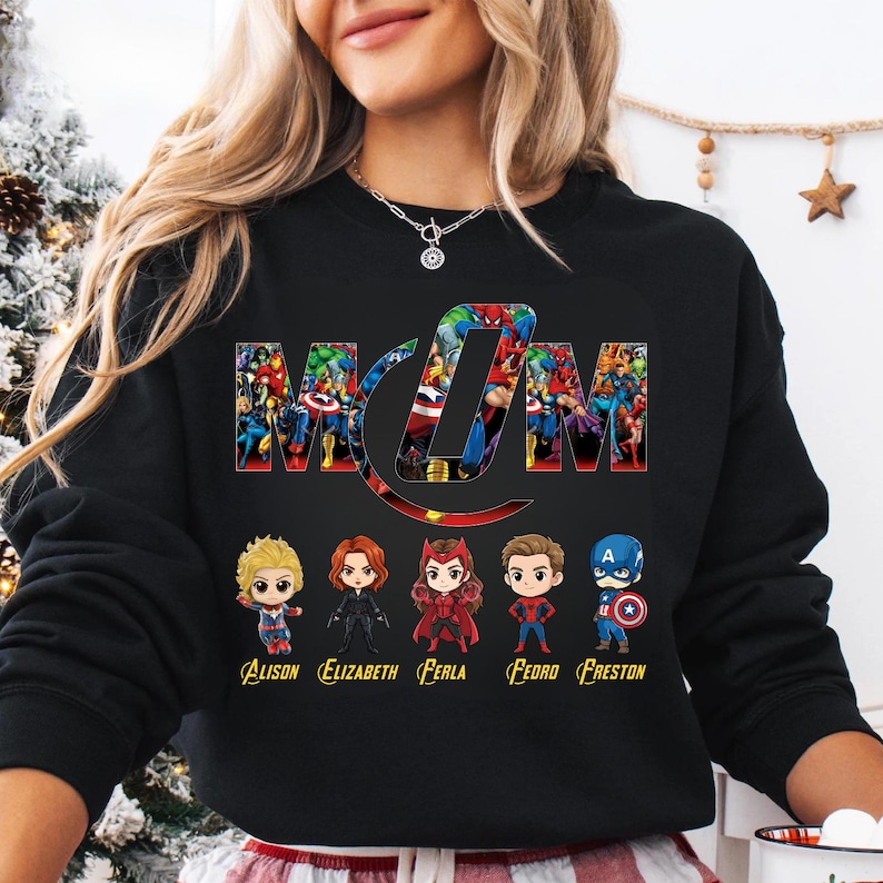 Peut inclure: Un sweat-shirt noir &agrave; manches longues avec le mot MOM en grandes lettres remplies de divers personnages de super-h&eacute;ros. Sous le texte se trouvent cinq figurines de super-h&eacute;ros style chibi avec les noms Alison, Elizabeth, Ferla, Fedro et Freston.