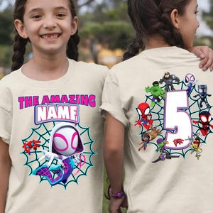 Op de afbeelding: Beige t-shirt met een Spider-Gwen-afbeelding, een webontwerp en de tekst "THE AMAZING NAME". De achterkant van het shirt toont het nummer 5 omringd door superhelden.