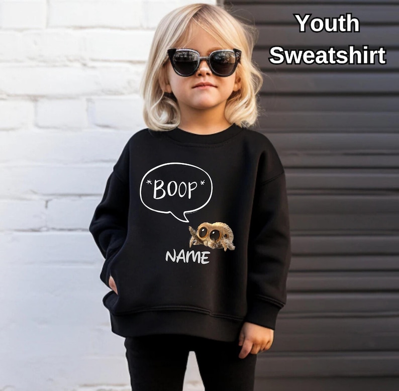 Puede incluir: Sudadera negra para j&oacute;venes con un bocadillo blanco que dice "*BOOP*" y una ara&ntilde;a de dibujos animados. Debajo de la ara&ntilde;a est&aacute; la palabra "NAME". La sudadera la lleva un ni&ntilde;o con gafas de sol.