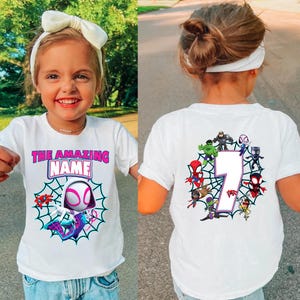 Op de afbeelding: Wit T-shirt met een Spider-Gwen ontwerp en de tekst "THE AMAZING NAME" in roze en blauw. De achterkant van het shirt toont het nummer 7 omringd door Spider-Man personages. Een leuk en kleurrijk ontwerp voor kinderen.