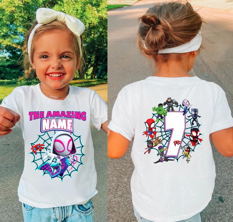 Op de afbeelding: Wit T-shirt met een Spider-Gwen ontwerp en de tekst "THE AMAZING NAME" in roze en blauw. De achterkant van het shirt toont het nummer 7 omringd door Spider-Man personages. Een leuk en kleurrijk ontwerp voor kinderen.