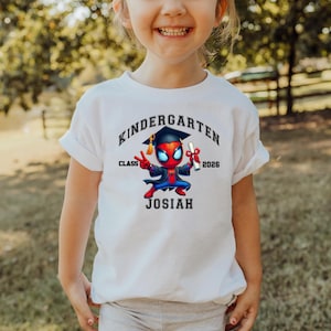 Puede incluir: Una camiseta blanca con un gr&aacute;fico de un superh&eacute;roe de dibujos animados con birrete y toga de graduaci&oacute;n, sosteniendo un diploma. El texto dice KINDERGARTEN, CLASS 2026 y JOSIAH en letras negras rodeando la ilustraci&oacute;n del personaje.