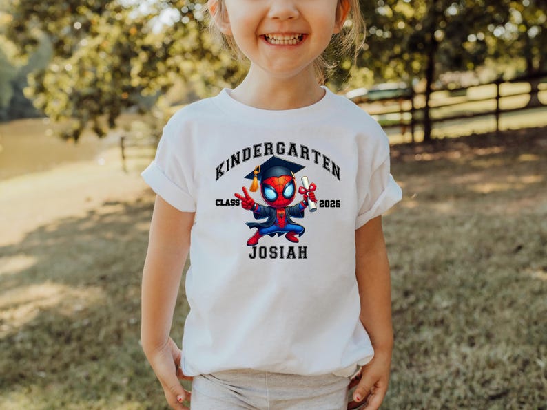 Puede incluir: Una camiseta blanca con un gr&aacute;fico de un superh&eacute;roe de dibujos animados con birrete y toga de graduaci&oacute;n, sosteniendo un diploma. El texto dice KINDERGARTEN, CLASS 2026 y JOSIAH en letras negras rodeando la ilustraci&oacute;n del personaje.