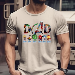 Peut inclure: Un t-shirt beige &agrave; manches courtes arborant le mot DAD en grandes lettres majuscules remplies d'images de super-h&eacute;ros. Sous le texte se trouvent six petites figurines de super-h&eacute;ros, chacune au-dessus du texte bleu Name.