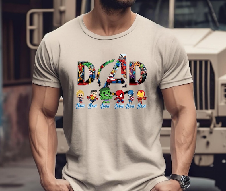 Peut inclure: Un t-shirt beige &agrave; manches courtes arborant le mot DAD en grandes lettres majuscules remplies d'images de super-h&eacute;ros. Sous le texte se trouvent six petites figurines de super-h&eacute;ros, chacune au-dessus du texte bleu Name.