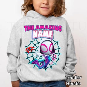 Op de afbeelding: Een lichtgrijze hoodie voor peuters met een cartoonontwerp van Spider-Gwen. Het ontwerp bevat een spinnenweb, de woorden "THE AMAZING NAME" in roze en blauw en de tekst "Toddler Hoodie".