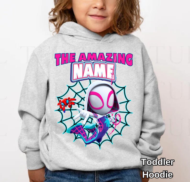 Op de afbeelding: Een lichtgrijze hoodie voor peuters met een cartoonontwerp van Spider-Gwen. Het ontwerp bevat een spinnenweb, de woorden "THE AMAZING NAME" in roze en blauw en de tekst "Toddler Hoodie".