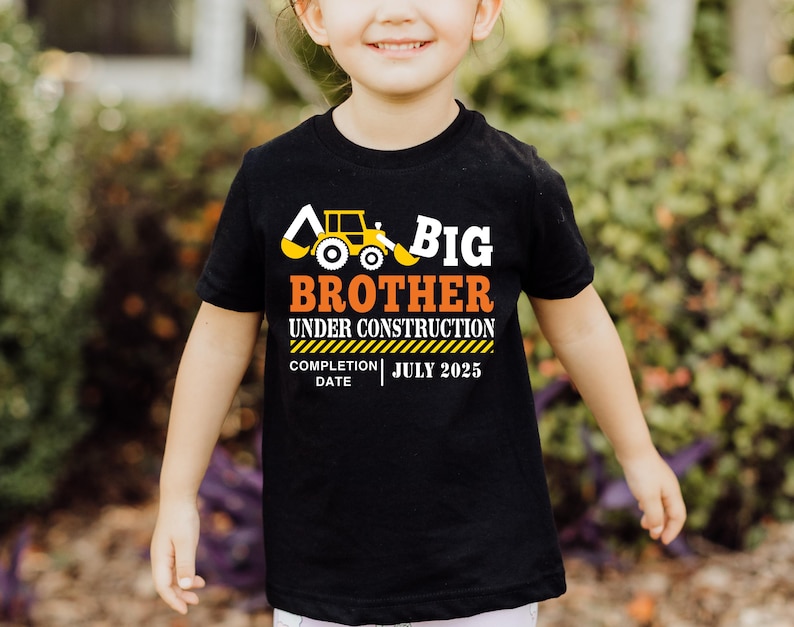 May include: Black t-shirt with the text "BIG BROTHER UNDER CONSTRUCTION" in white and orange, featuring a yellow construction vehicle graphic. The completion date is July 2025. The shirt is worn by a child.