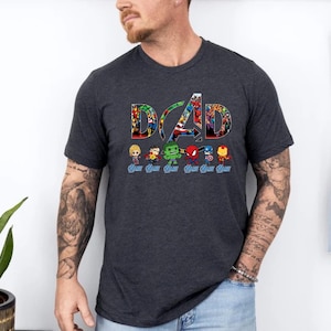 Peut inclure: Un t-shirt gris anthracite &agrave; manches courtes avec le mot DAD en grandes lettres remplies d'un collage de super-h&eacute;ros color&eacute;s. Sous le texte se trouvent six petites figurines de super-h&eacute;ros, chacune avec le mot DAD &eacute;crit en bleu en dessous.