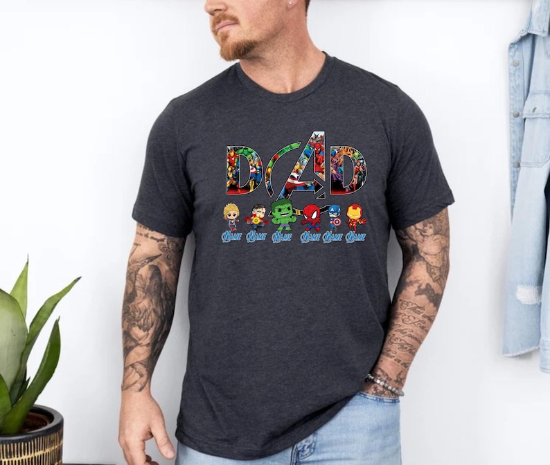 Peut inclure: Un t-shirt gris anthracite &agrave; manches courtes avec le mot DAD en grandes lettres remplies d'un collage de super-h&eacute;ros color&eacute;s. Sous le texte se trouvent six petites figurines de super-h&eacute;ros, chacune avec le mot DAD &eacute;crit en bleu en dessous.