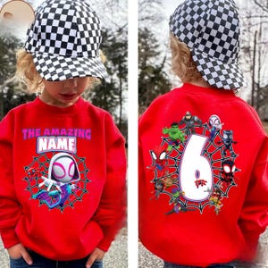 Op de afbeelding: Een rode trui met een Spider-Gwen-ontwerp en de tekst "THE AMAZING NAME". De achterkant van de trui toont het cijfer 6, omringd door verschillende superhelden. Het kind draagt een zwart-wit geblokte baseballcap.