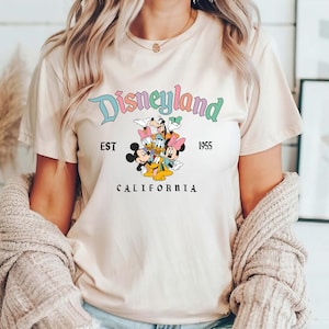 Puede incluir: Una camiseta beige claro con un gr&aacute;fico de Disneyland colorido. El gr&aacute;fico presenta a Mickey Mouse, Minnie Mouse, Donald Duck, Goofy y Pluto. El texto "Disneyland" est&aacute; en una fuente de arco&iacute;ris con las palabras "Est. 1955" y "California" debajo del gr&aacute;fico.