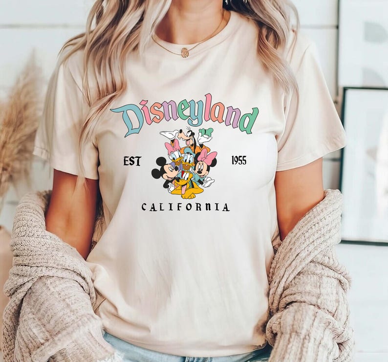 Puede incluir: Una camiseta beige claro con un gr&aacute;fico de Disneyland colorido. El gr&aacute;fico presenta a Mickey Mouse, Minnie Mouse, Donald Duck, Goofy y Pluto. El texto "Disneyland" est&aacute; en una fuente de arco&iacute;ris con las palabras "Est. 1955" y "California" debajo del gr&aacute;fico.