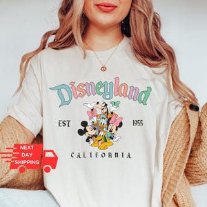 Puede incluir: Camiseta blanca con un logotipo de Disneyland colorido, con Mickey Mouse, Minnie Mouse, Donald Duck, Goofy y Pluto. El texto dice "Disneyland Est 1955 California".