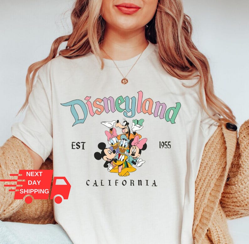 Puede incluir: Camiseta blanca con un logotipo de Disneyland colorido, con Mickey Mouse, Minnie Mouse, Donald Duck, Goofy y Pluto. El texto dice "Disneyland Est 1955 California".